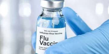 A gloved hand is holding a 5 mL vial labeled "Influenza Virus Vaccine, Flu Vaccine." A syringe needle is being inserted into the vial, drawing up the liquid vaccine. The background is blurred, highlighting the focus on the vaccine vial and syringe, reminiscent of various Moderna stock images. | FintechZoom