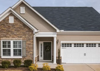 A single-story property with a blend of beige siding and stone facade, featuring a gabled roof with dark shingles. The front includes a well-maintained lawn, shrubs near the entrance, a large garage with white doors, and shuttered windows. The sky is partly cloudy. | FintechZoom