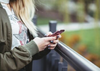 A person with blonde hair is standing outside, holding and looking at a pink smartphone, perhaps browsing some entertainment. They are wearing a green jacket and a light-colored shirt with a design on it. Their nails are painted greyish-blue. A blurred outdoor scene is in the background. | FintechZoom