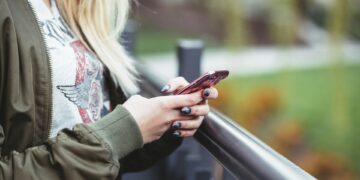 A person with blonde hair is standing outside, holding and looking at a pink smartphone, perhaps browsing some entertainment. They are wearing a green jacket and a light-colored shirt with a design on it. Their nails are painted greyish-blue. A blurred outdoor scene is in the background. | FintechZoom