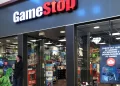 The image shows the entrance of a GameStop store in a mall. The store is brightly lit, with colorful game-related merchandise displayed inside. A man, perhaps keeping an eye on the GME share price, is standing outside the store, looking at his phone while holding a takeaway drink. | FintechZoom