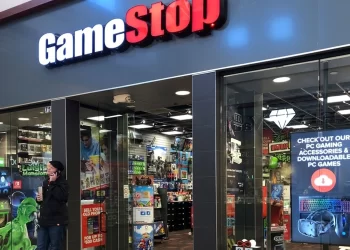 The image shows the entrance of a GameStop store in a mall. The store is brightly lit, with colorful game-related merchandise displayed inside. A man, perhaps keeping an eye on the GME share price, is standing outside the store, looking at his phone while holding a takeaway drink. | FintechZoom