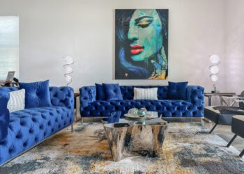A modern living room features two blue tufted sofas crafted from sustainable furniture materials with white pillows. A glass coffee table with chrome accents stands in the center, beneath a colorful painting of a woman's face. Two contemporary floor lamps flank the sofas, and a patterned rug covers the floor. | FintechZoom