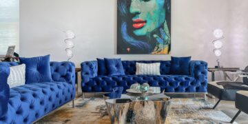 A modern living room features two blue tufted sofas crafted from sustainable furniture materials with white pillows. A glass coffee table with chrome accents stands in the center, beneath a colorful painting of a woman's face. Two contemporary floor lamps flank the sofas, and a patterned rug covers the floor. | FintechZoom