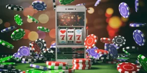 A smartphone displaying a slot machine with "777" on the screen sits among scattered poker chips of various colors, including red, blue, black, green, and white. The background is blurred with colorful bokeh lights, creating a festive casino-like atmosphere perfect for playing online slots. | FintechZoom