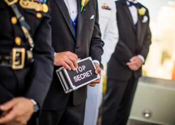 A group of men wearing military uniforms and formal suits stand in a line. One man in the center, cloaked in pseudonymity, is holding a small metallic briefcase labeled "TOP SECRET." The background is blurred, focusing attention on the men and the briefcase. | FintechZoom