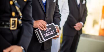 A group of men wearing military uniforms and formal suits stand in a line. One man in the center, cloaked in pseudonymity, is holding a small metallic briefcase labeled "TOP SECRET." The background is blurred, focusing attention on the men and the briefcase. | FintechZoom