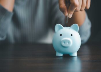 A person wearing a gray top is placing a coin into a blue piggy bank on a dark surface, perhaps inspired by searching for the best CD rates in Florida. The focus is on the piggy bank and the person's hand, while the background is blurred. | FintechZoom