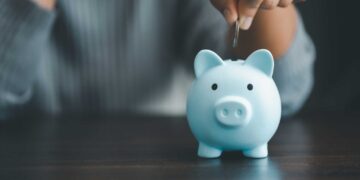 A person wearing a gray top is placing a coin into a blue piggy bank on a dark surface, perhaps inspired by searching for the best CD rates in Florida. The focus is on the piggy bank and the person's hand, while the background is blurred. | FintechZoom