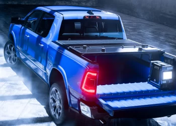 A sleek, blue pickup truck with its tailgate down and bed partially illuminated by the surrounding blue and white lights is showcased in a modern, industrial studio setting. The truck has stylish rims and a Worksport stock tonneau cover, enhancing the overall futuristic and clean atmosphere. | FintechZoom