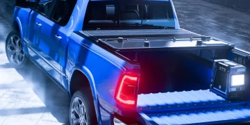 A sleek, blue pickup truck with its tailgate down and bed partially illuminated by the surrounding blue and white lights is showcased in a modern, industrial studio setting. The truck has stylish rims and a Worksport stock tonneau cover, enhancing the overall futuristic and clean atmosphere. | FintechZoom