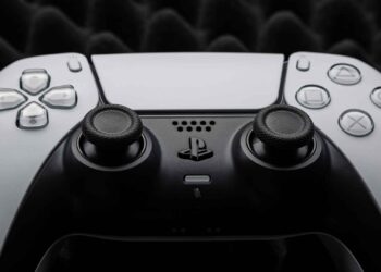 A close-up photo of a white PlayStation 5 controller, showcasing the directional pad, analog sticks, and face buttons including the triangle, circle, cross, and square icons. The background features a textured, dark foam material perfect for any gaming setup. | FintechZoom