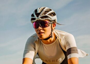 A cyclist wearing a white helmet, reflective sunglasses, and a fitted white jersey rides outdoors under a clear sky. The helmet has the logo "Kask," and the jersey has the logo "Rapha" on its sleeve. With his gear featuring some of the best sunglasses for cycling, he appears focused and determined. | FintechZoom