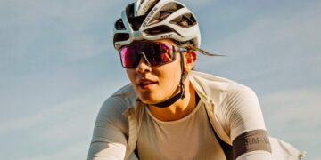 A cyclist wearing a white helmet, reflective sunglasses, and a fitted white jersey rides outdoors under a clear sky. The helmet has the logo "Kask," and the jersey has the logo "Rapha" on its sleeve. With his gear featuring some of the best sunglasses for cycling, he appears focused and determined. | FintechZoom