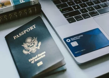 Close-up of a desk with a United States passport, the best travel credit card—Chase Sapphire Preferred—and an open laptop. Part of a book is visible in the corner, titled "ROUTE." The passport is slightly worn, while the card and laptop are in good condition. | FintechZoom