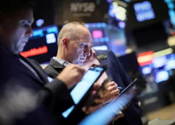 Several people, focused on their screens, are engaged in monitoring and trading activities on the floor of a stock exchange. Digital price boards in the background display fluctuating market values, prominently featuring the Dow Jones Industrial Average today, contributing to the dynamic and fast-paced environment. | FintechZoom