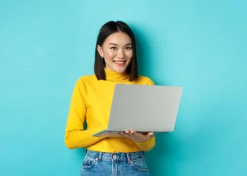 A woman with shoulder-length dark hair wearing a yellow sweater and blue jeans smiles while holding an open laptop displaying the best CD rates. She stands against a solid teal background. | FintechZoom