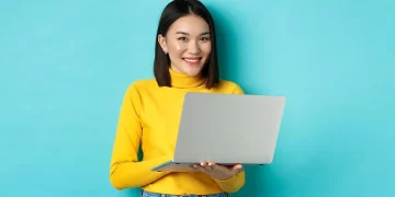 A woman with shoulder-length dark hair wearing a yellow sweater and blue jeans smiles while holding an open laptop displaying the best CD rates. She stands against a solid teal background. | FintechZoom