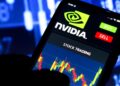 A smartphone screen displays the NVIDIA logo with "BUY" and "SELL" buttons. Below it, a stock trading chart with colorful candlesticks is shown. In the background, a blurred image of stock market data on a monitor provides context to the live action. | FintechZoom
