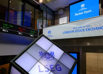 A view inside the London Stock Exchange shows digital displays. One screen reads "Welcome to the London Stock Exchange," another displays stock details in red and green. The LSEG logo is prominently featured on a tilted cube-shaped display in the foreground, with FTSE 100 highlights capturing attention. | FintechZoom