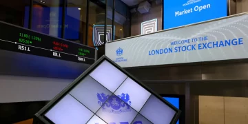 A view inside the London Stock Exchange shows digital displays. One screen reads "Welcome to the London Stock Exchange," another displays stock details in red and green. The LSEG logo is prominently featured on a tilted cube-shaped display in the foreground, with FTSE 100 highlights capturing attention. | FintechZoom