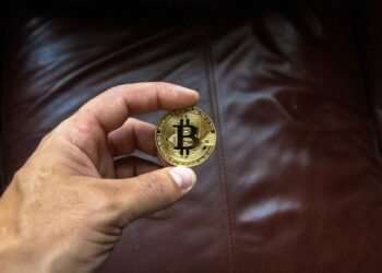 A hand holding a gold-colored Bitcoin coin. The background features a dark brown leather surface, partially visible. The Bitcoin logo and some text are engraved on the coin, highlighting the prominence of cryptocurrencies in modern finance. | FintechZoom
