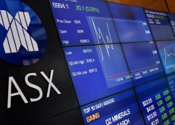 Image of the Australian Securities Exchange (ASX) trading board displaying various stock prices and market indices. The large blue ASX logo is prominent on the left, while various financial data and charts are shown on the digital screens to the right. | FintechZoom