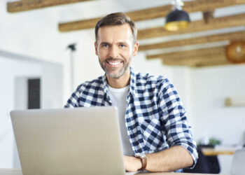 A smiling man with a beard and mustache is sitting at a table using a laptop, possibly reviewing a PDF. He is wearing a blue and white checkered shirt. The room has exposed wooden beams on the ceiling, white walls, and modern decor. The ambiance appears bright and inviting. | FintechZoom