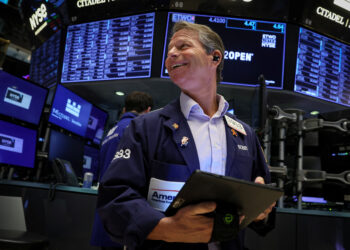 A trader at the New York Stock Exchange, wearing a dark blue jacket with various badges, smiles while holding a tablet. Multiple screens with stock information and the NYSE logo are visible in the background. | FintechZoom