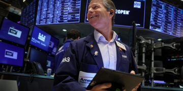 A trader at the New York Stock Exchange, wearing a dark blue jacket with various badges, smiles while holding a tablet. Multiple screens with stock information and the NYSE logo are visible in the background. | FintechZoom