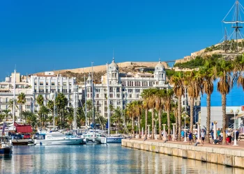 A picturesque marina scene with various boats docked in calm waters, surrounded by palm trees. In the background, white historic buildings with ornate facades stand against a clear blue sky, showcasing Alicante Real Estate. Hills and a partially visible sailboat add depth to the view. | FintechZoom