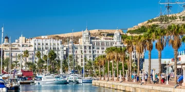 A picturesque marina scene with various boats docked in calm waters, surrounded by palm trees. In the background, white historic buildings with ornate facades stand against a clear blue sky, showcasing Alicante Real Estate. Hills and a partially visible sailboat add depth to the view. | FintechZoom