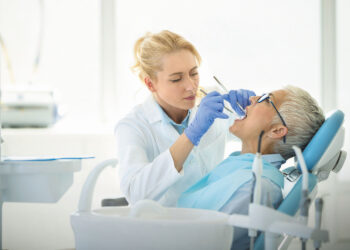A dentist wearing a white coat and blue gloves examines a patient’s teeth using dental instruments. The patient, wearing a blue bib, is reclining in a dental chair with their mouth open during the dental care examination. The examination room is brightly lit. | FintechZoom