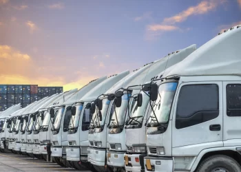 A row of white commercial trucks is parked in an orderly line under a colorful sky at sunset. Shipping containers are stacked in the background. The trucks appear ready for transport, mindful of common DOT violations, emphasizing logistics and transportation themes. | FintechZoom
