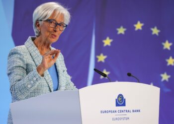 A person with short white hair and glasses is speaking at a podium with the European Central Bank logo, discussing the ECB's plan to cut rates. They are wearing a light blue suit and gesturing with their hand. The background features the European Union flag with yellow stars on a blue field. | FintechZoom