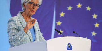 A person with short white hair and glasses is speaking at a podium with the European Central Bank logo, discussing the ECB's plan to cut rates. They are wearing a light blue suit and gesturing with their hand. The background features the European Union flag with yellow stars on a blue field. | FintechZoom