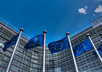 A row of tall flagpoles displaying blue flags with yellow stars representing the European Union stands in front of a modern glass building with a curved facade, reminiscent of corporate structures housing FTSE 100 companies. The sky is clear blue with a few small clouds. | FintechZoom