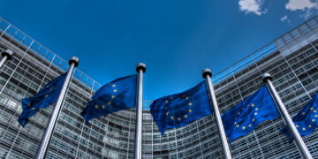 A row of tall flagpoles displaying blue flags with yellow stars representing the European Union stands in front of a modern glass building with a curved facade, reminiscent of corporate structures housing FTSE 100 companies. The sky is clear blue with a few small clouds. | FintechZoom