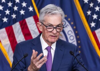 A man with gray hair and glasses, wearing a blue suit and purple tie, speaks at a podium about the latest Federal Reserve decision. Behind him are two American flags and a blue curtain with the seal of the U.S. Federal Reserve, partially visible. | FintechZoom