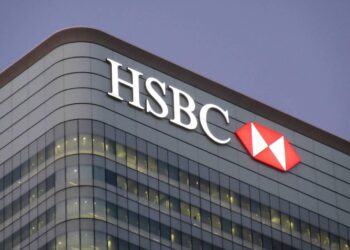 A modern glass building featuring the HSBC logo, with white letters and a red and white hexagonal emblem, prominently displayed at the top. The building has rows of lit windows and is set against a dusky evening sky, subtly hinting at recent UK banking glitches. | FintechZoom