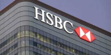 A modern glass building featuring the HSBC logo, with white letters and a red and white hexagonal emblem, prominently displayed at the top. The building has rows of lit windows and is set against a dusky evening sky, subtly hinting at recent UK banking glitches. | FintechZoom