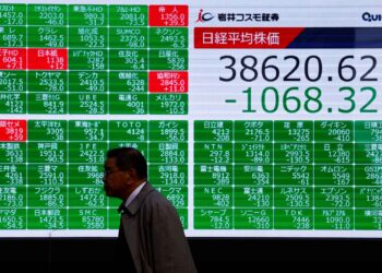 A man walks past a large electronic stock board showing the Nikkei 225 down by 1068.32 points, with a closing value of 38620.62. The screen displays multiple individual stock prices, most highlighted in green, and the information is primarily in Japanese. | FintechZoom
