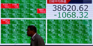 A man walks past a large electronic stock board showing the Nikkei 225 down by 1068.32 points, with a closing value of 38620.62. The screen displays multiple individual stock prices, most highlighted in green, and the information is primarily in Japanese. | FintechZoom