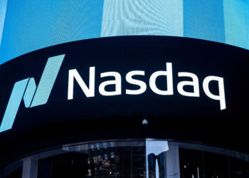 A large digital display shows the Nasdaq logo, featuring a stylized "N" and the text "Nasdaq" in white against a black background. Located on the side of a building, its surface reflects surrounding architecture while nearby, another screen highlights movements in the S&P 500. | FintechZoom