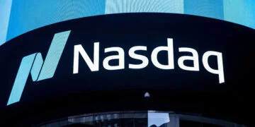 A large digital display shows the Nasdaq logo, featuring a stylized "N" and the text "Nasdaq" in white against a black background. Located on the side of a building, its surface reflects surrounding architecture while nearby, another screen highlights movements in the S&P 500. | FintechZoom