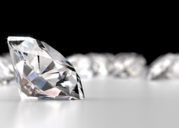 Close-up image of a single polished lab-grown diamond in sharp focus, sitting on a reflective surface. Several blurred diamonds are visible in the background against a black backdrop, creating a contrast that highlights the brilliance and facets of the featured diamond. | FintechZoom