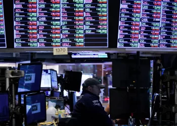 A stock trader sits at a desk surrounded by multiple monitors displaying stock market information. Large electronic boards overhead show various stock tickers and financial data in different colors, illustrating Market Analysis Today and indicating market trends and activity on a busy trading floor. | FintechZoom