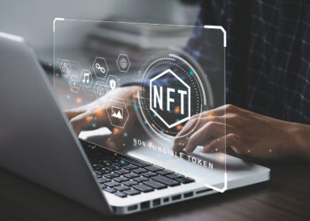 A person is typing on a laptop with a floating digital graphic in front of the screen showing the text "NFT" and "Non-Fungible Token." The graphic includes icons of various digital assets, indicating NFT-related content or transactions, showcasing the world of NFTs. | FintechZoom
