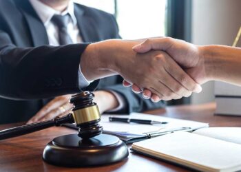 Two individuals are shaking hands across a wooden table. One person, possibly a personal injury attorney, is wearing a suit, suggesting a professional setting. On the table, there is a gavel, an open notebook, and legal documents. | FintechZoom