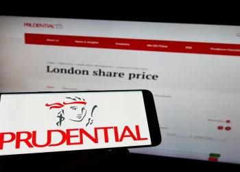 A smartphone displays the Prudential logo in front of a computer screen showing the Prudential website with the "London share price" section open. The setup highlights Prudential's branding and a segment of their online financial information, emphasizing their position within the FTSE 100. | FintechZoom